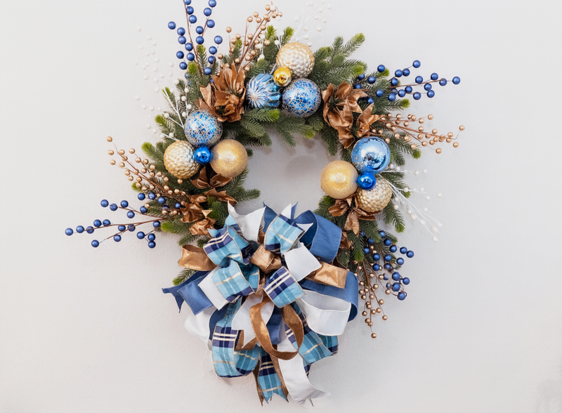 Decorative wreath comprised of blue plaid, ivory, and sparkling metallic gold ribbons, blue and gold ornaments, blue and gold berry picks, on a neutral background