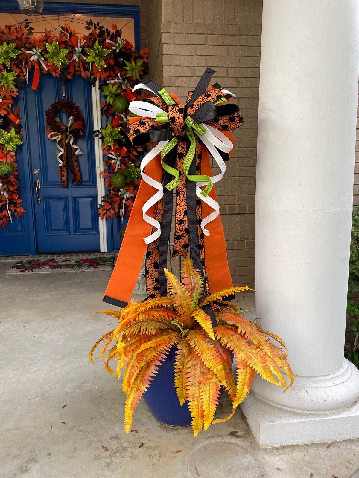 Close up view of a bow comprised of 4" Orange w/ black backing double-fused dupion ribbon 2.5"Orange w/ black flocked spiders and flocked black backing ribbon 1.5" d. stevens Black pleated metallic ribbon 1" White pleated metallic ribbon 1" Lime green ribbon with lime rhinestone center hanging from trellis in a large planter with an orange fern.
