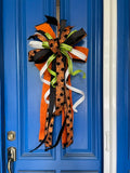 Close up view of a bow comprised of 4" Orange w/ black backing double-fused dupion ribbon 2.5"Orange w/ black flocked spiders and flocked black backing ribbon 1.5" d. stevens Black pleated metallic ribbon 1" White pleated metallic ribbon 1" Lime green ribbon with lime rhinestone center hanging from a blue door