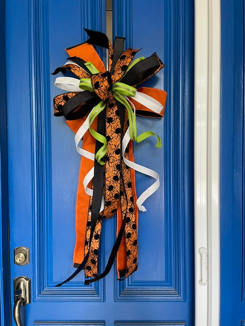Close up view of a bow comprised of 4" Orange w/ black backing double-fused dupion ribbon 2.5"Orange w/ black flocked spiders and flocked black backing ribbon 1.5" d. stevens Black pleated metallic ribbon 1" White pleated metallic ribbon 1" Lime green ribbon with lime rhinestone center hanging from a blue door