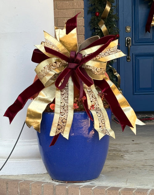 Large decorative bow for wreaths or door hangers comprised of a 4" d. stevens heavily rhinestone embellished, gold ribbon, a 2.5" burgundy velvet ribbon, and a 1" burgundy velvet ribbon on a trellis in a planter with a a blue front door in the background