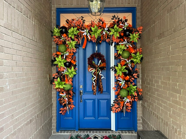 Photo of oversized blue door with a very large garland wrapped around the top and sides. There is also a wreath with a large bow. Colors include orange, black, white, and lime green.