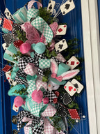 Close up image of the right side of a large decorative teardrop shaped door swag comprised of a 2.5" Aqua blue and white checker board patterned ribbon, a 2.5" Pink and white checker board patterned ribbon, and a 1.5" Black and white checker board patterned ribbon, accented with a tea pot and tea cup, a rabbit, top hat, clock, and playing cards on a blue door