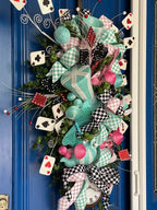 Close up image of the left side of a large decorative teardrop shaped door swag comprised of a 2.5" Aqua blue and white checker board patterned ribbon, a 2.5" Pink and white checker board patterned ribbon, and a 1.5" Black and white checker board patterned ribbon, accented with a tea pot and tea cup, a rabbit, top hat, clock, and playing cards on a blue door