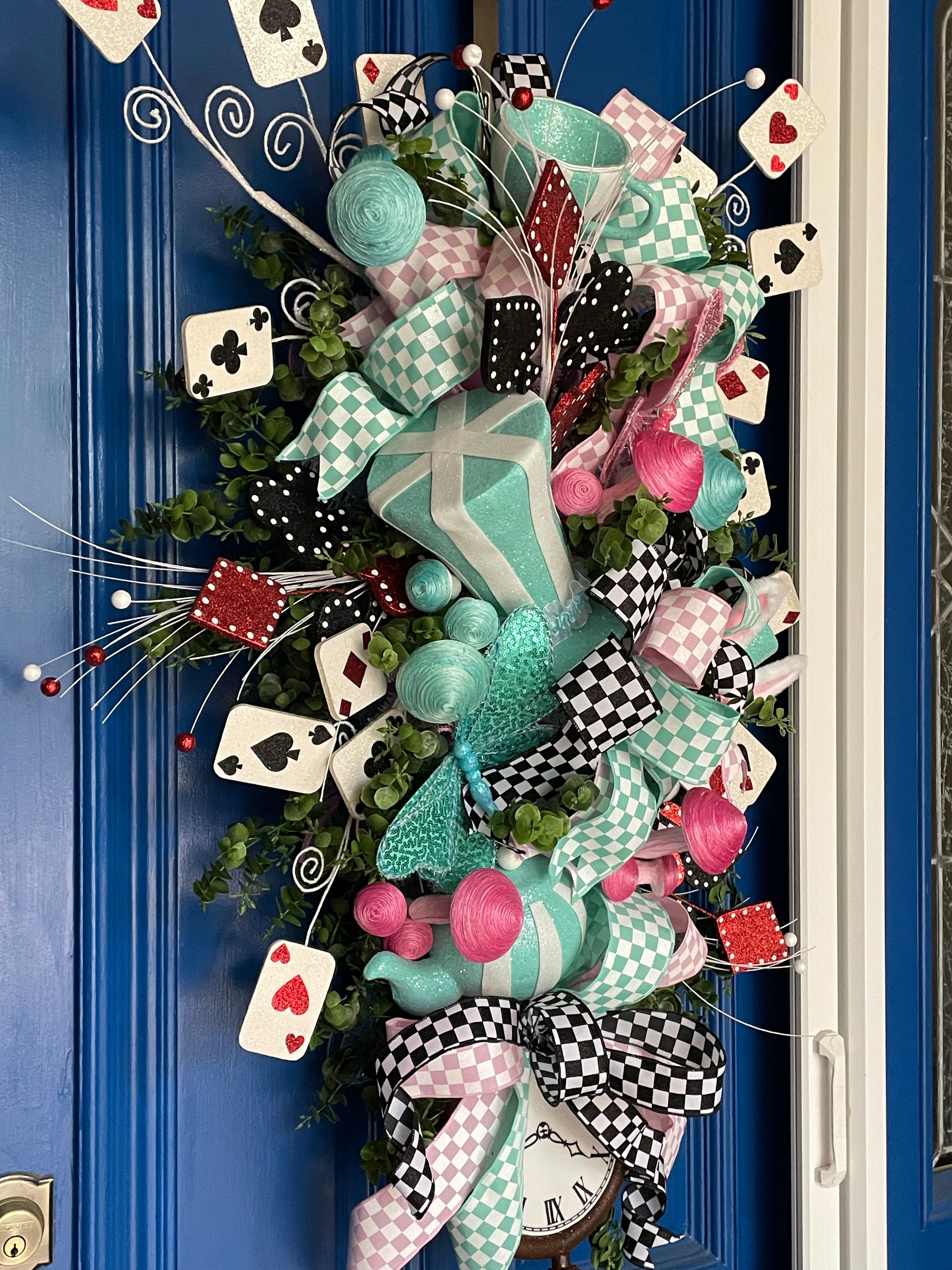 Close up image of the left side of a large decorative teardrop shaped door swag comprised of a 2.5" Aqua blue and white checker board patterned ribbon, a 2.5" Pink and white checker board patterned ribbon, and a 1.5" Black and white checker board patterned ribbon, accented with a tea pot and tea cup, a rabbit, top hat, clock, and playing cards on a blue door