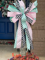 Close up of a large decorative bow for wreaths or door hangers comprised of a 2.5" Aqua blue and white checker board patterned ribbon, a 2.5" Pink and white checker board patterned ribbon, and a1.5" Black and white checker board patterned ribbon on a trellis in a large blue planter in on a front porch with a blue door and matching door swag in the background
