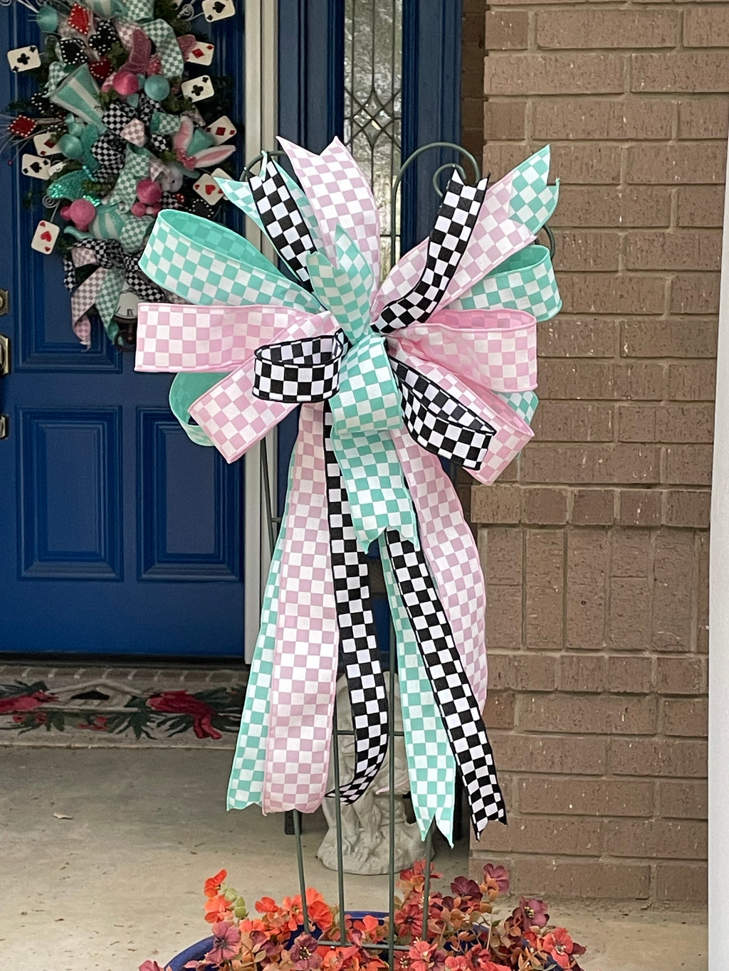 Large decorative bow for wreaths or door hangers comprised of a 2.5" Aqua blue and white checker board patterned ribbon, a 2.5" Pink and white checker board patterned ribbon, and a1.5" Black and white checker board patterned ribbon on a trellis in a large blue planter in on a front porch with a matching door swag on a blue door in the background