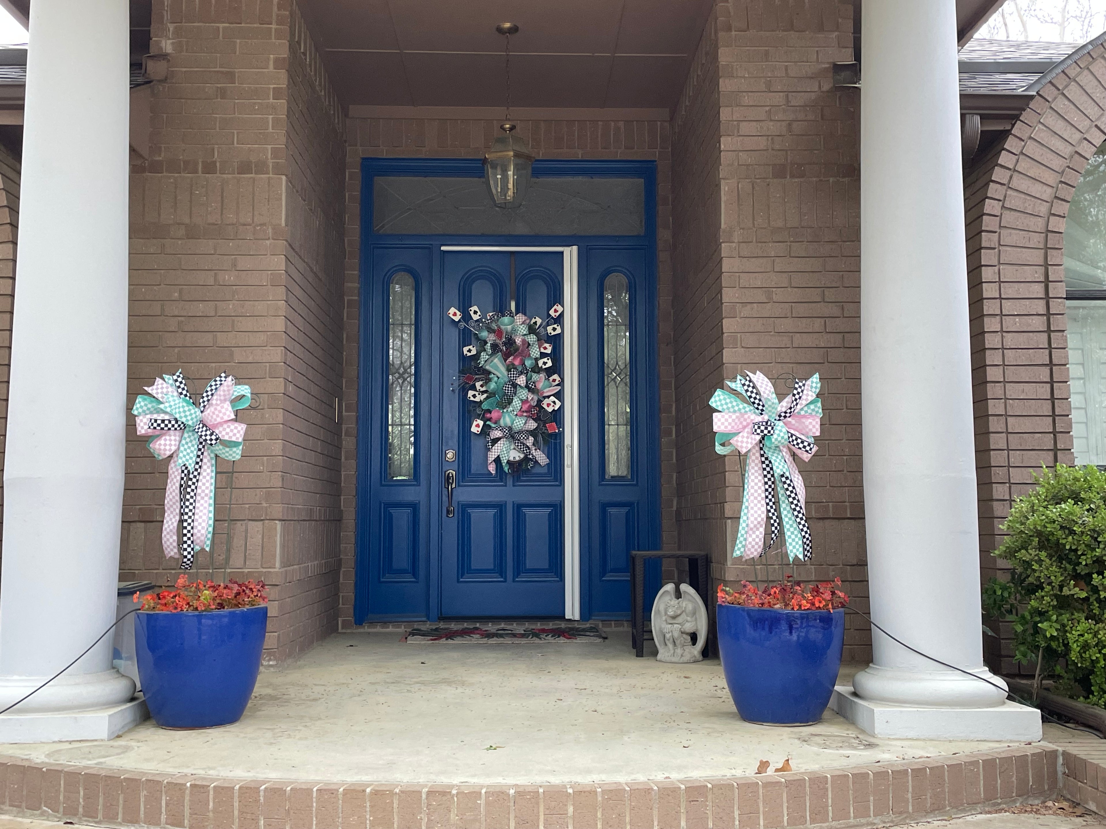 Wide shot of a large decorative bows for wreaths or door hangers comprised of a 2.5" Aqua blue and white checker board patterned ribbon, a 2.5" Pink and white checker board patterned ribbon, and a1.5" Black and white checker board patterned ribbon on a trellis in a large blue planters in on a front porch with a blue door and matching door swag in the background