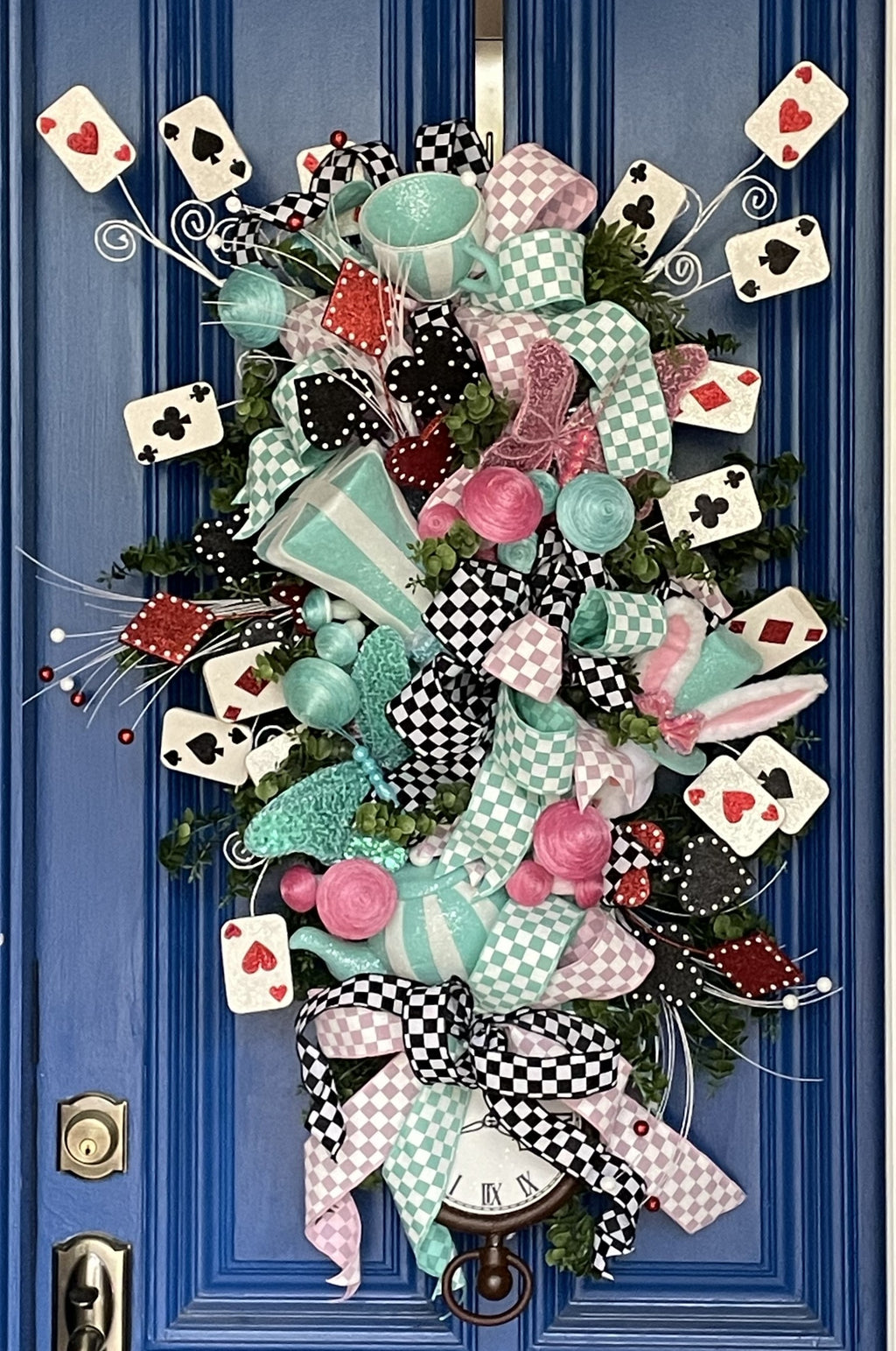 Large decorative teardrop shaped door swag comprised of a 2.5" Aqua blue and white checker board patterned ribbon, a 2.5" Pink and white checker board patterned ribbon, and a 1.5" Black and white checker board patterned ribbon, accented with a tea pot and tea cup, a rabbit, top hat, clock, and playing cards on a blue door