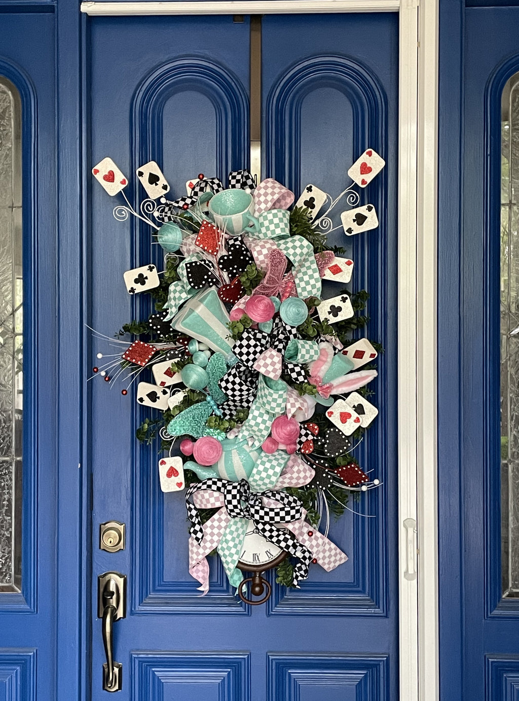 Close up video of a a large decorative teardrop shaped door swag comprised of a 2.5" Aqua blue and white checker board patterned ribbon, a 2.5" Pink and white checker board patterned ribbon, and a 1.5" Black and white checker board patterned ribbon, accented with a tea pot and tea cup, a rabbit, top hat, clock, and playing cards on a blue door