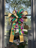 Large decorative bow for wreaths or door hangers comprised of a Farrisilk 4" gold velvet, a Farrisilk 1" heavily rhinestoned purple metallic ribbon, a Farrisilk 1" pleated; metallic green ribbon, and a Farrisilk 2.5" gold velvet ribbon center accent on a glass door