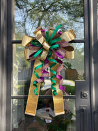 Large decorative bow for wreaths or door hangers comprised of a Farrisilk 4" gold velvet, a Farrisilk 1" heavily rhinestoned purple metallic ribbon, a Farrisilk 1" pleated; metallic green ribbon, and a Farrisilk 2.5" gold velvet ribbon center accent on a glass door