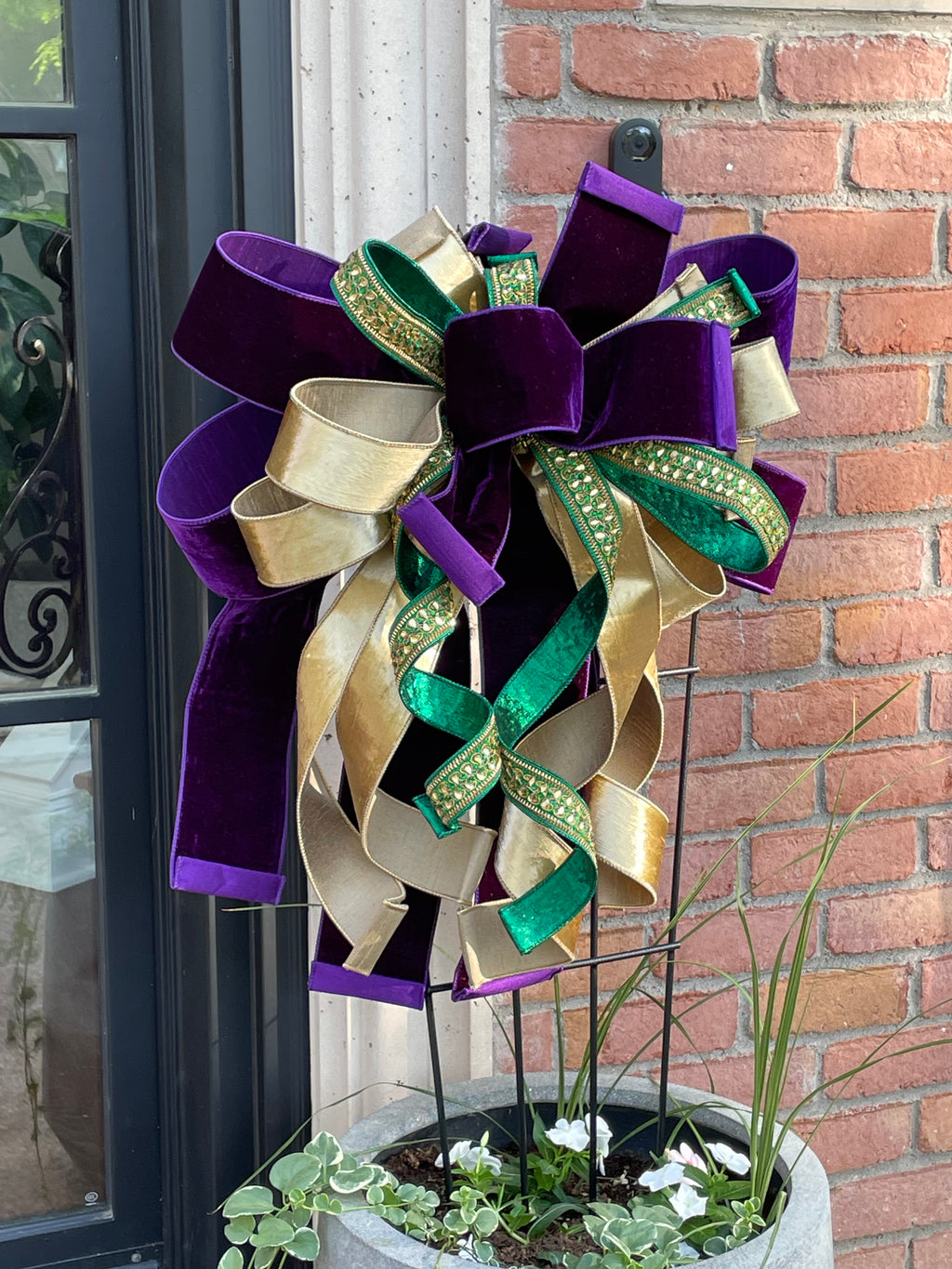 Large decorative bow for wreaths or door hangers comprised of a 4" Farrisilk purple velvet ribbon, a 2.5" Farrisilk gold velvet ribbon center, and a 1" Farrisilk heavily rhinestoned green metallic ribbon on a trellis in a large planter