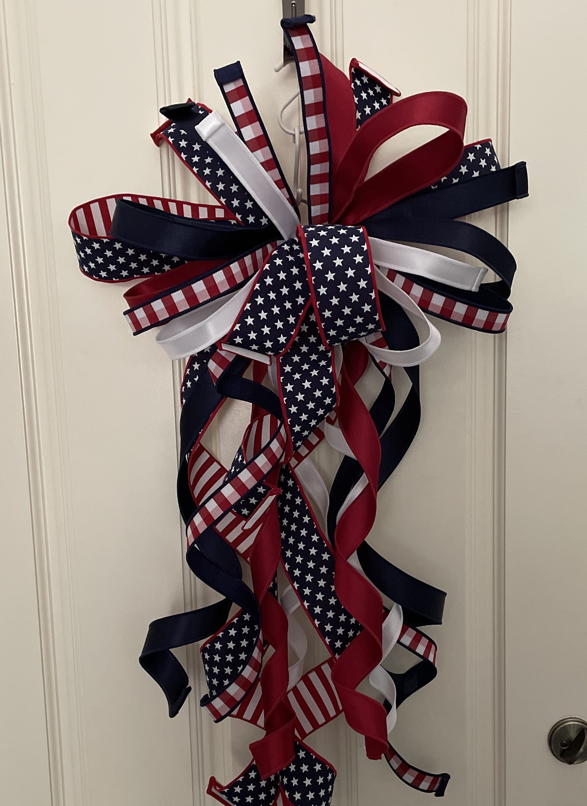 Large decorative bow for wreaths or door hangers comprised of a 1" d. stevens red and white check ribbon with navy blue dupion backing, a 2.5" stars and stripes ribbon, a 1.5" red satin ribbon, a 1.5" navy satin ribbon, and a 1" white satin ribbon on a neutral background. Shown with tails curled.