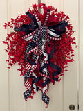 Decorative wreath with dainty red flowers and a bow comprised of a 2.5" Stars and Stripes Ribbon, a 1" red, white and navy blue checked ribbon, a 1.5" red satin ribbon, a 1.5" navy satin ribbon, and a 1" white satin ribbon on a neutral colored door.