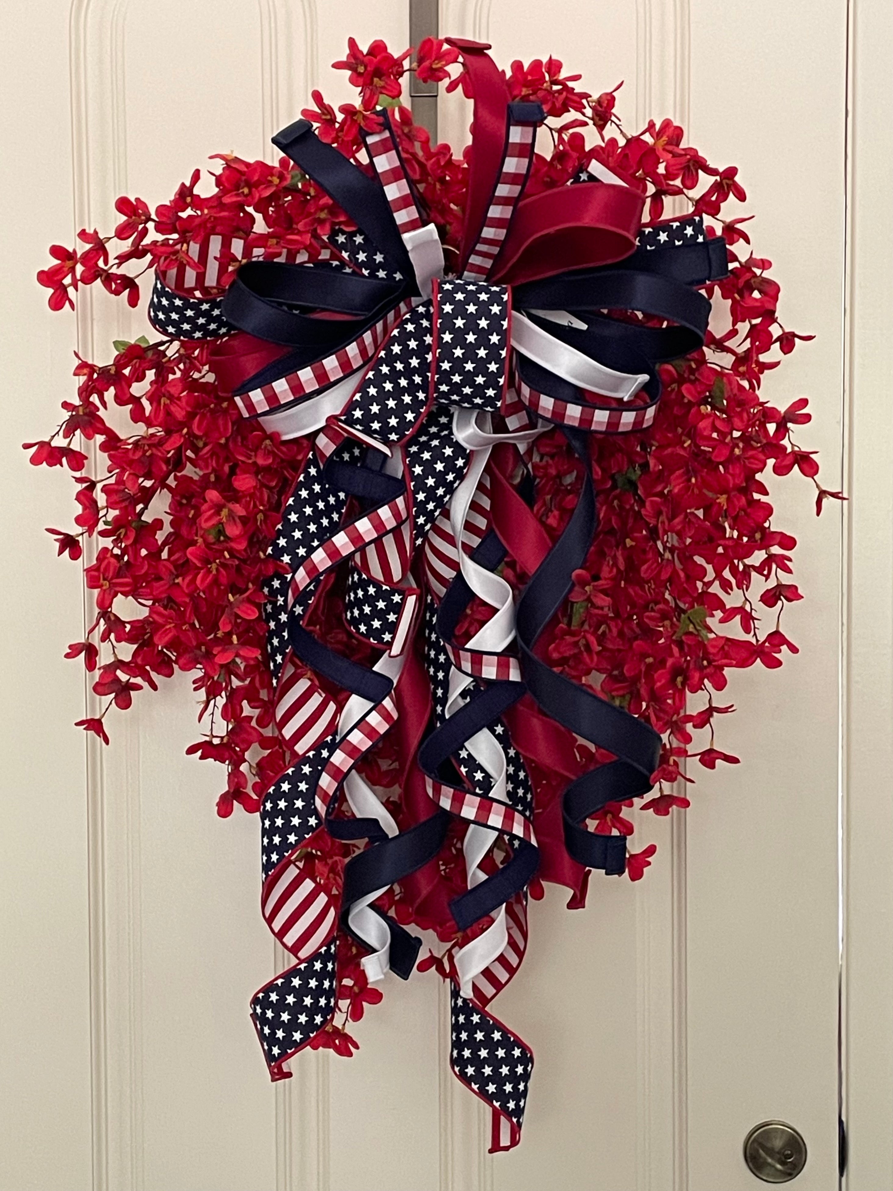 Decorative wreath with dainty red flowers and a bow comprised of a 2.5" Stars and Stripes Ribbon, a 1" red, white and navy blue checked ribbon, a 1.5" red satin ribbon, a 1.5" navy satin ribbon, and a 1" white satin ribbon on a neutral colored door.