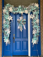 Large decorative garland comprised of ice blue ribbon with glittered white snowflakes, ice blue satin ribbon, large styrofoam snowflakes, white faux fur flowers, glittery ice blue berry picks, glittery ice blue snowflakes, on a blue door with a coordinating large decorative bow for wreaths or door hangers hanging in the center of the door