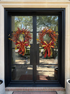 A pair of designer bows compromised of 2/5" Burgundy taffeta ribbon, 1" gold dupion ribbon, and 1.5" dark orange ribbon attached at the bottom of a fall berry wreath which is mounted onto  glass doors.