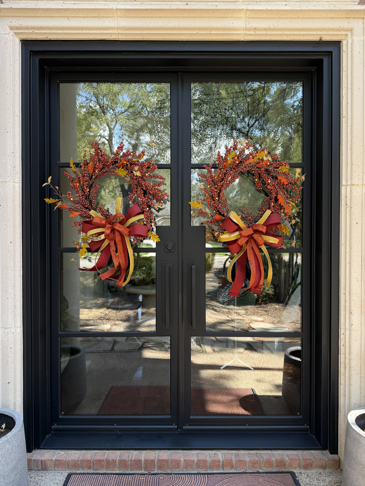 A pair of designer bows compromised of 2/5" Burgundy taffeta ribbon, 1" gold dupion ribbon, and 1.5" dark orange ribbon attached at the bottom of a fall berry wreath which is mounted onto  glass doors.