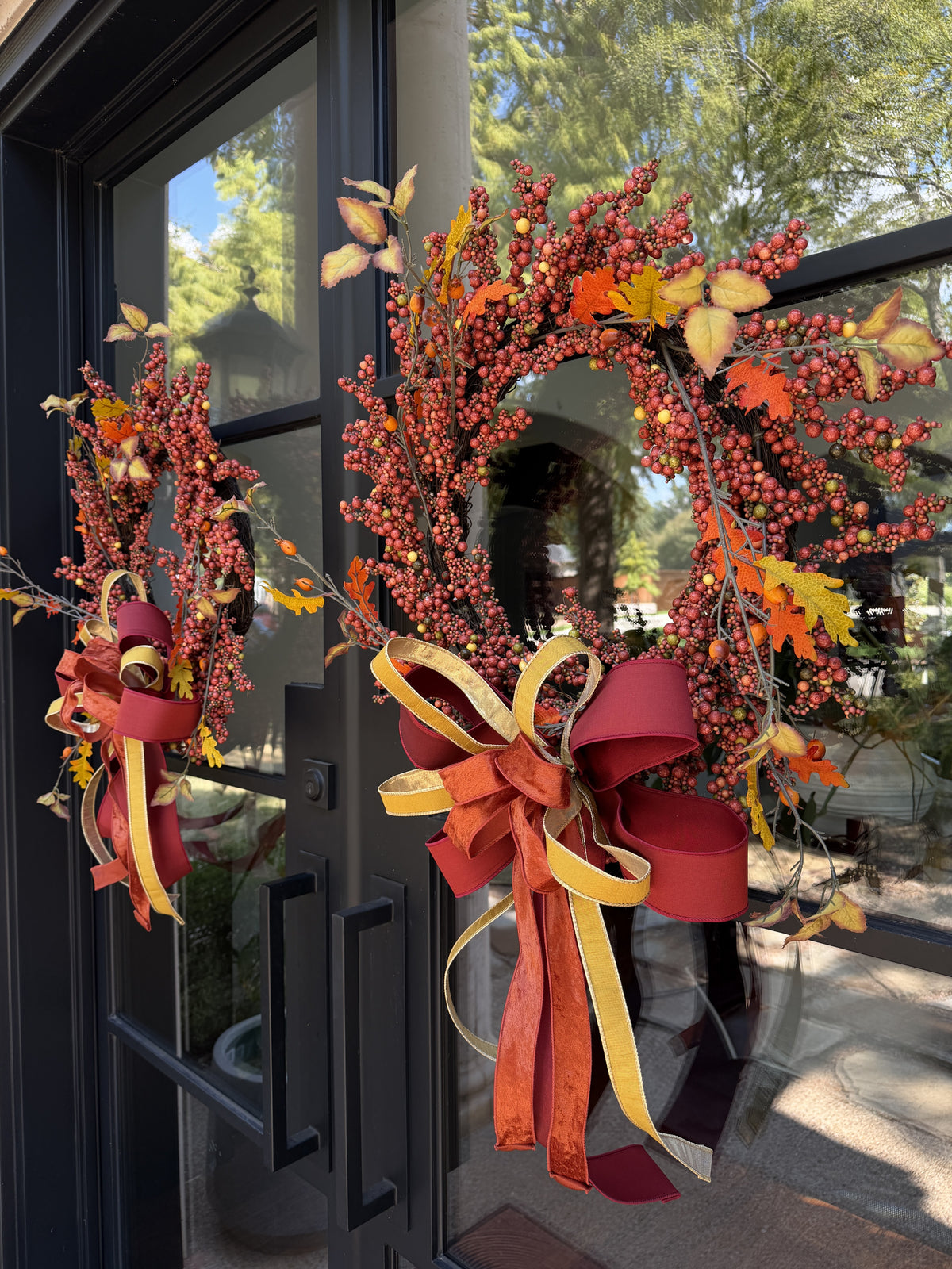 Side view of a pair of designer bows compromised of 2/5" Burgundy taffeta ribbon, 1" gold dupion ribbon, and 1.5" dark orange ribbon attached at the bottom of a fall berry wreath which is mounted onto  glass doors.