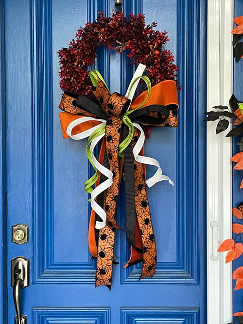 Picture of a Red and orange artificial boxwood wreath with an oversized box comprised of orange, black, orange with black spiders, white, and lime green ribbons. The bow is hanging on a blue door.