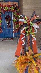 Close up video of a bow comprised of 4" Orange w/ black backing double-fused dupion ribbon 2.5"Orange w/ black flocked spiders and flocked black backing ribbon 1.5" d. stevens Black pleated metallic ribbon 1" White pleated metallic ribbon 1" Lime green ribbon with lime rhinestone center hanging from a trellis in a large blue planter - in front of a doorway decorated with coordinating wreath and garlands.