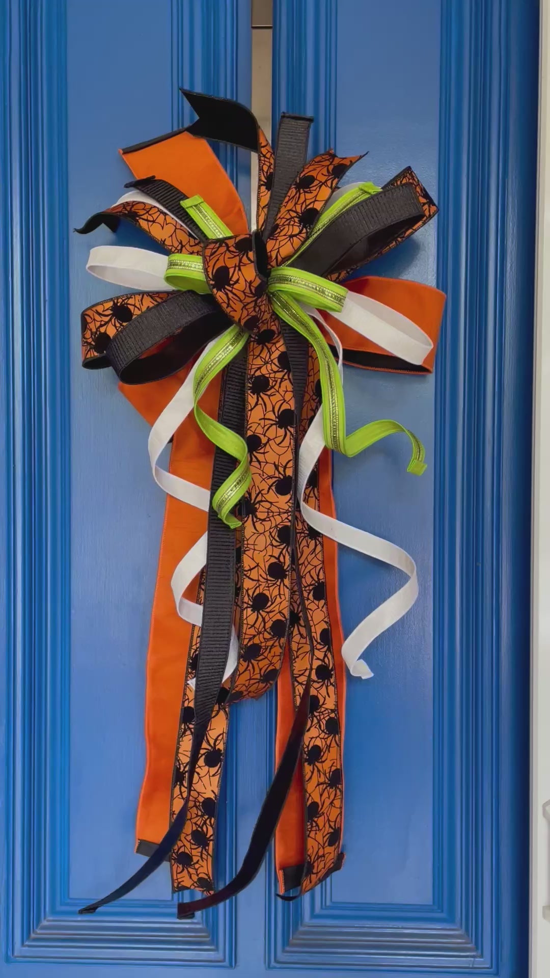 Close up video of a bow comprised of 4" Orange w/ black backing double-fused dupion ribbon 2.5"Orange w/ black flocked spiders and flocked black backing ribbon 1.5" d. stevens Black pleated metallic ribbon 1" White pleated metallic ribbon 1" Lime green ribbon with lime rhinestone center hanging from a blue door