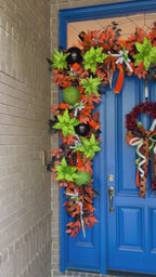 Video of an oversized blue door with a very large garland wrapped around the top and sides. There is also a wreath with a large bow. Colors include orange, black, white, and lime green.