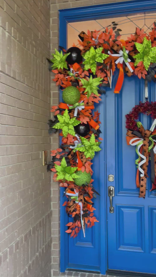 Video of an oversized blue door with a very large garland wrapped around the top and sides. There is also a wreath with a large bow. Colors include orange, black, white, and lime green.