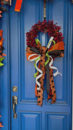 Video of a red and orange artificial boxwood wreath with an oversized box comprised of orange, black, orange with black spiders, white, and lime green ribbons. The bow is hanging on a blue door.