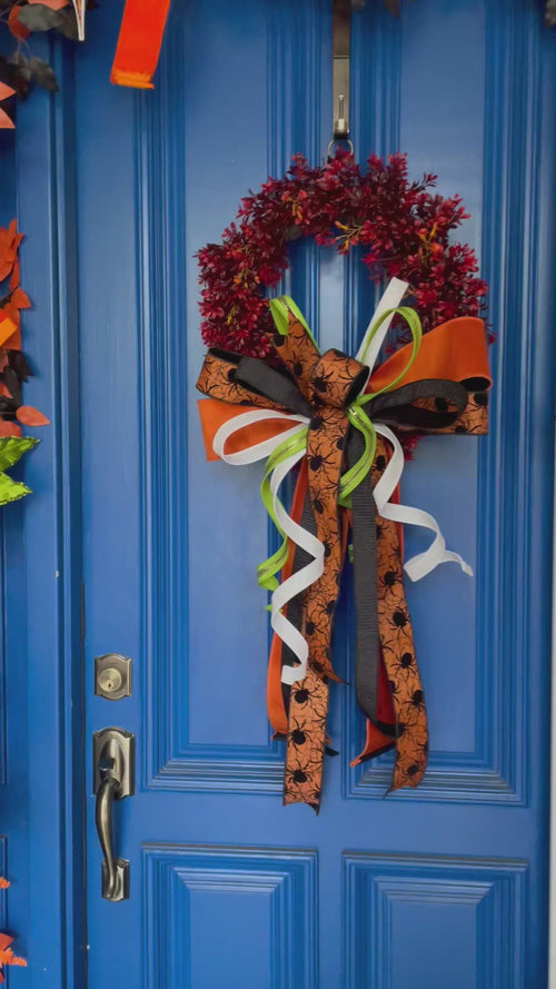 Video of a red and orange artificial boxwood wreath with an oversized box comprised of orange, black, orange with black spiders, white, and lime green ribbons. The bow is hanging on a blue door.