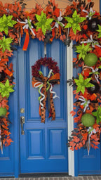 A 2nd video of an oversized blue door with a very large garland wrapped around the top and sides. There is also a wreath with a large bow. Colors include orange, black, white, and lime green.